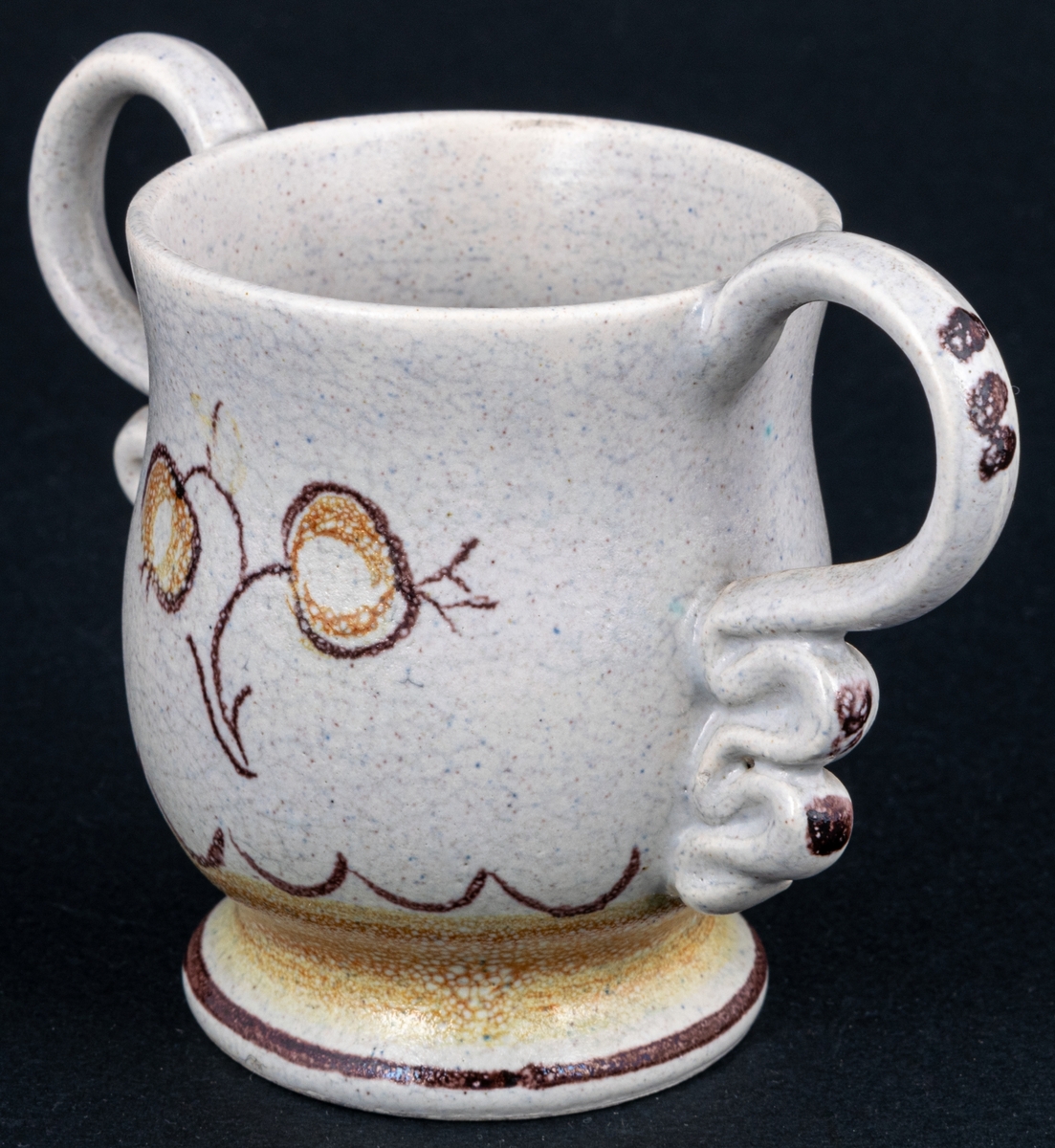 Bägare av lergods. Bo Fajans, design Eva Jancke­Björk. Grå S­svängd med stora hänklar, ljus- och mörkbruna partier nedtill samt dekor av en blomma.