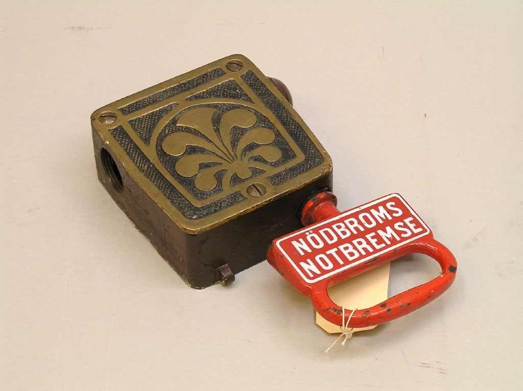 Nödbroms med handtag. Själva "dosan" är av mässing med dekor av en stiliserad växt.
Text på det röda handtaget: NÖDBROMS NOTBREMSE