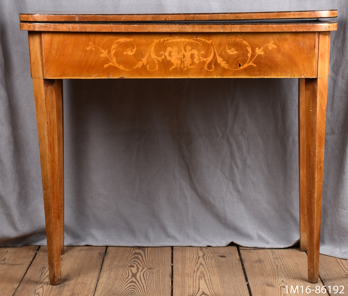 Spelbord av mahogny. På framsidan av sargen dekorationer i intarsia. Skivan, nedfällbar, är liksidig med avrundade hörn. Raka, nedåt avsmalnande ben. Se 1M16-86191.