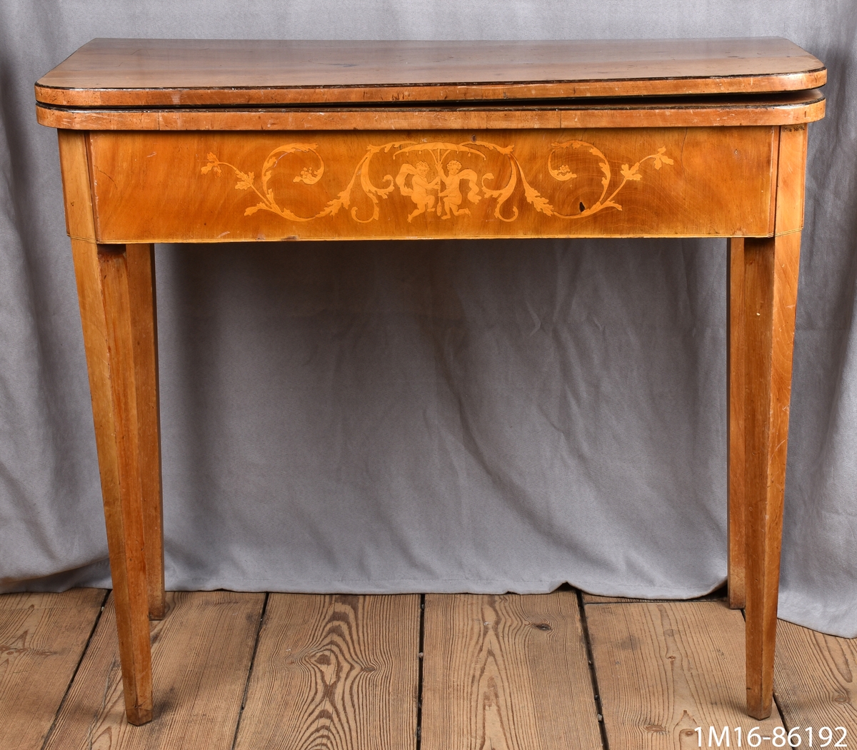 Spelbord av mahogny. På framsidan av sargen dekorationer i intarsia. Skivan, nedfällbar, är liksidig med avrundade hörn. Raka, nedåt avsmalnande ben. Se 1M16-86191.