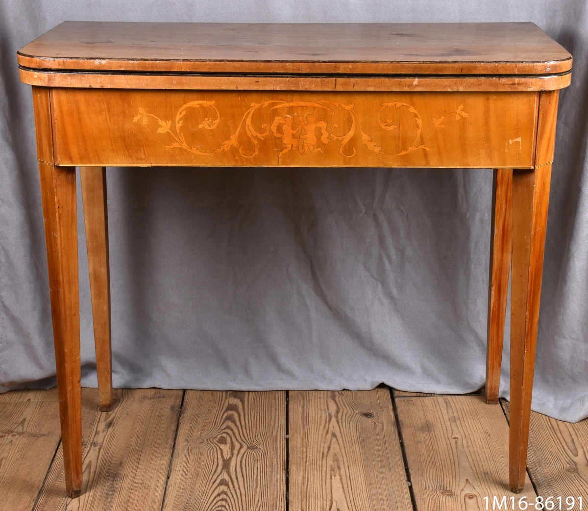 Spelbord av mahogny. På framsidan av sargen dekorationer i intarsia. Skivan, nedfällbar, är liksidig med avrundade hörn. Raka, nedåt avsmalnande ben.