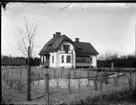 En okänd större putsad villa byggd i vinkel. Ett byggnadsår på väggen ovanför en balkong säger möjligen 1904. En nyanlagd trädgård med träd och buskar i förgrunden.