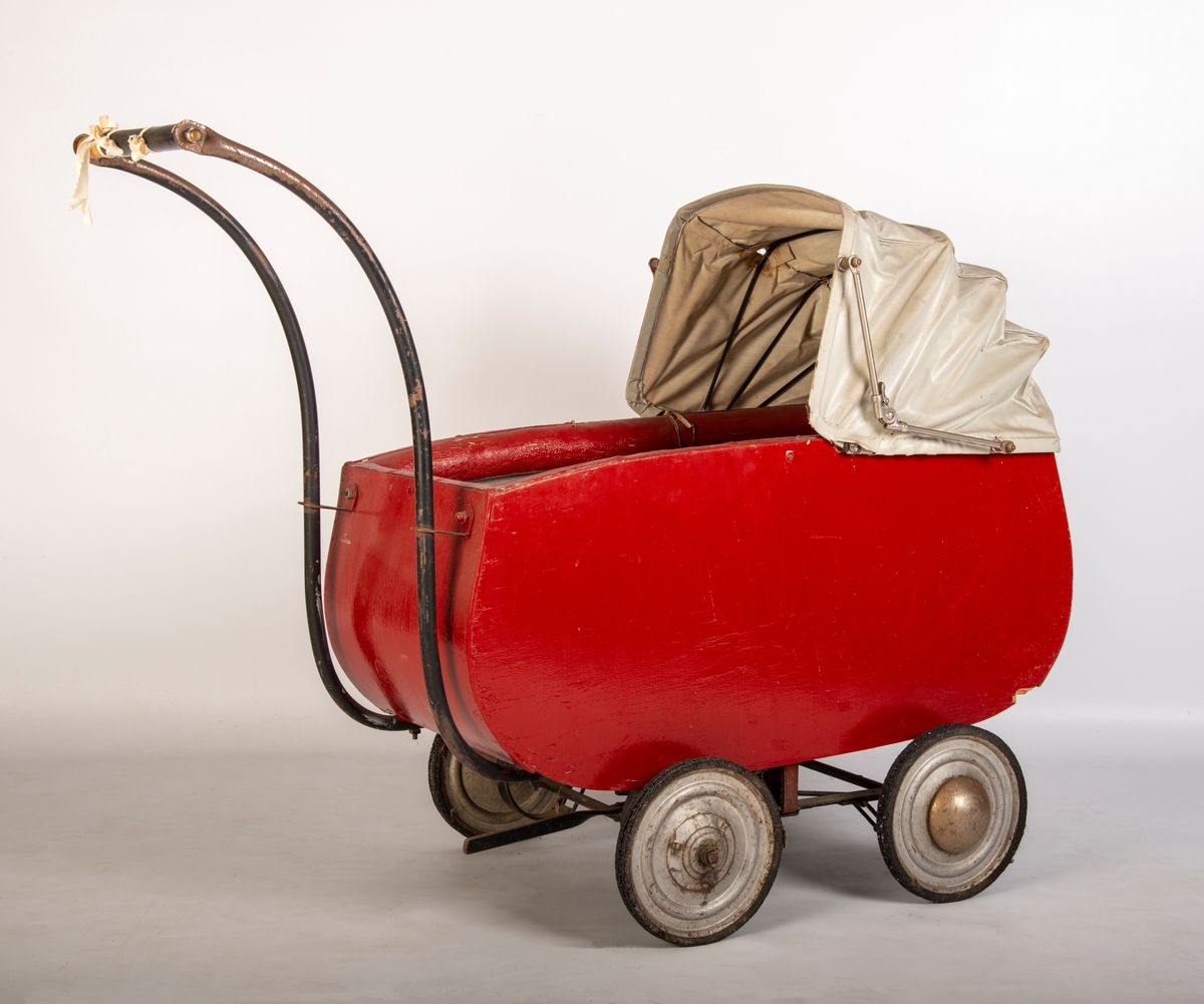 Rødmalt vogn med gråmalt innerside og lys grå kalesje av kunstlær. 4 metallhjul med nedslitt gummidekk. Håndtak av tre med svart plastlignende materiale som er sprøtt. Kalesje med ei flenge. Polstret karm. Mangler skrue i håndtak og kalesje.