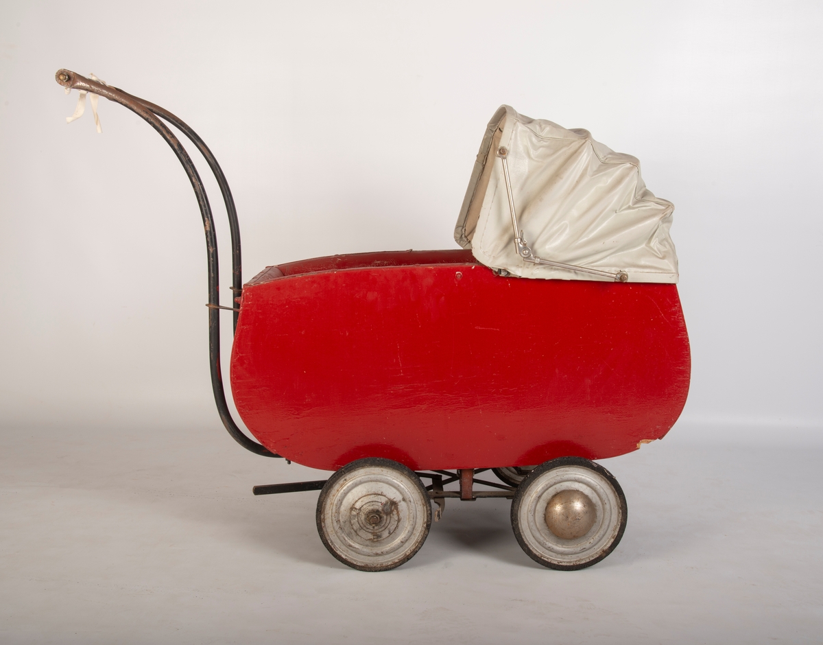Rødmalt vogn med gråmalt innerside og lys grå kalesje av kunstlær. 4 metallhjul med nedslitt gummidekk. Håndtak av tre med svart plastlignende materiale som er sprøtt. Kalesje med ei flenge. Polstret karm. Mangler skrue i håndtak og kalesje.