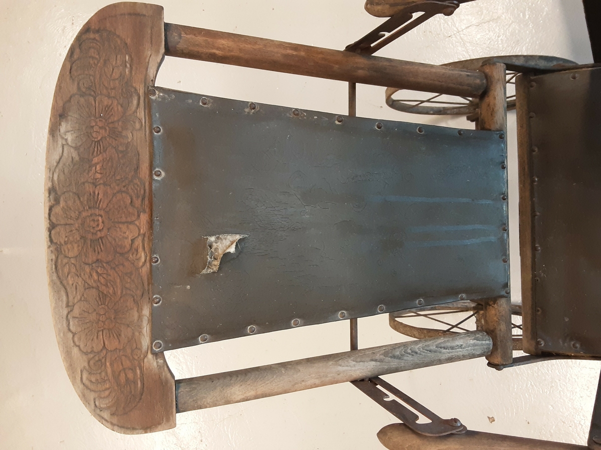 Ramme av tre med kunstlær i sete og rygg. Håndtak av jernrør. 4 hjul mangler dekk. Mangler stopper for fotbrett. Hull i ryggtrekket og mye rust. Setetrekket løsnet i den ene siden. noen enkle reparasjoner med ståltråd.