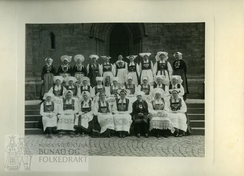 Samling av kvinner i ulike bunader frå Hordaland på ei steintrapp ufanfor ei kyrkje. Sjå baksida av biletet.