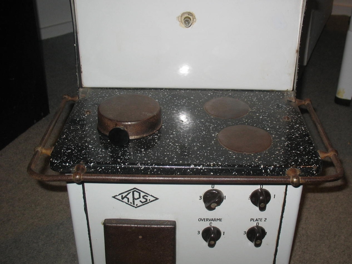 Miniatyrutgave av magasinkomfyr fra KPS. Jfr. BrM.16287.
Hvitemaljerte sider og front. Topplaten er sortemaljert med hvite prikker. Komfyren har 2 plater, 1 magasin og stekeovn. Den har fire funksjonsbrytere. En av bryterene er tilkoblet lyset på ryggplaten