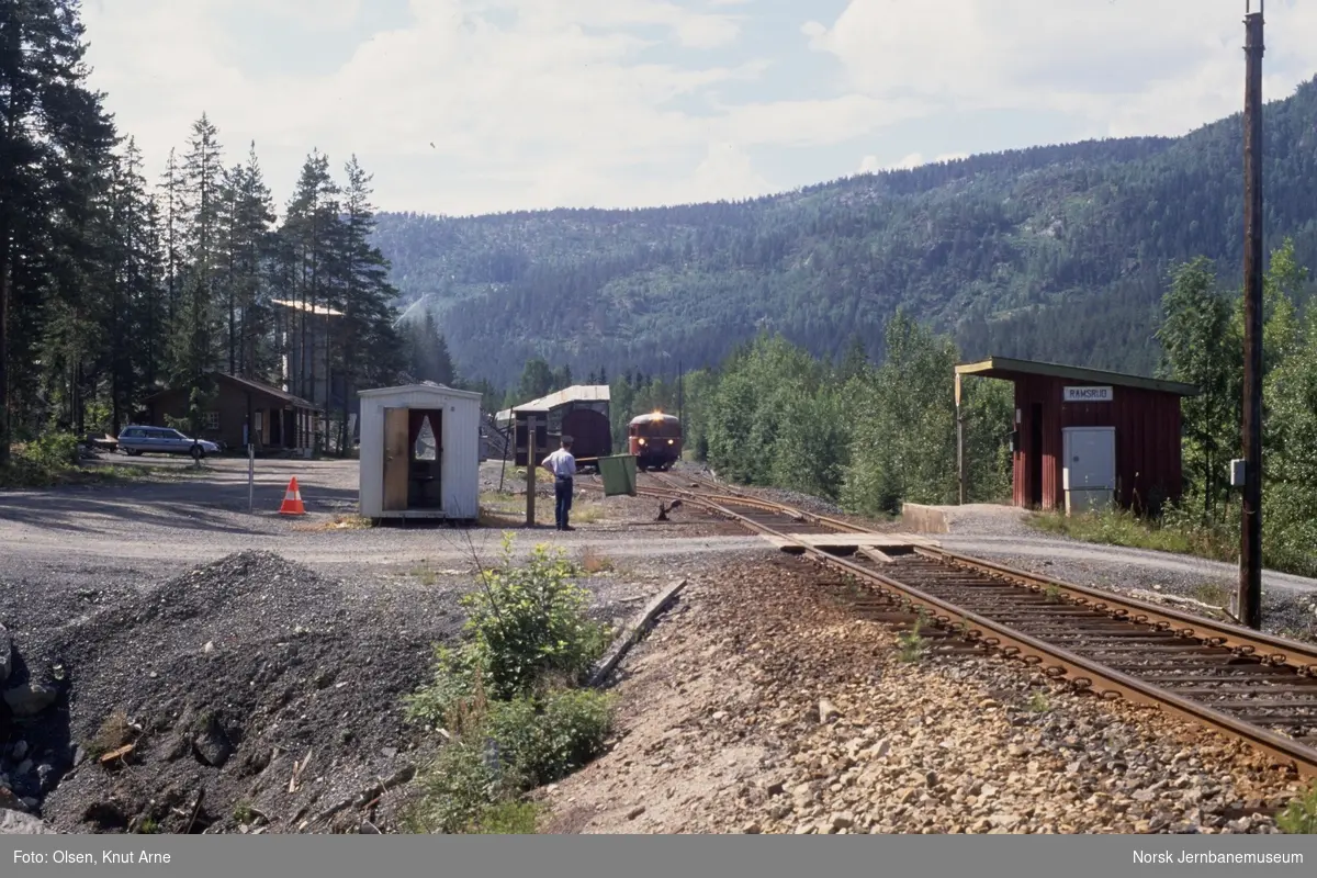 Ramsrud holdeplass er opprettet midlertidig stasjon med signalsted ved sidesporet, og togekspeditør Karsten Brodal viser grønt flagg. Denne løsningen ble benyttet ved arbeider på banen. I bakgrunnen dieselmotorvogn BM 86 25 med persontog fra Kongsberg til Rødberg