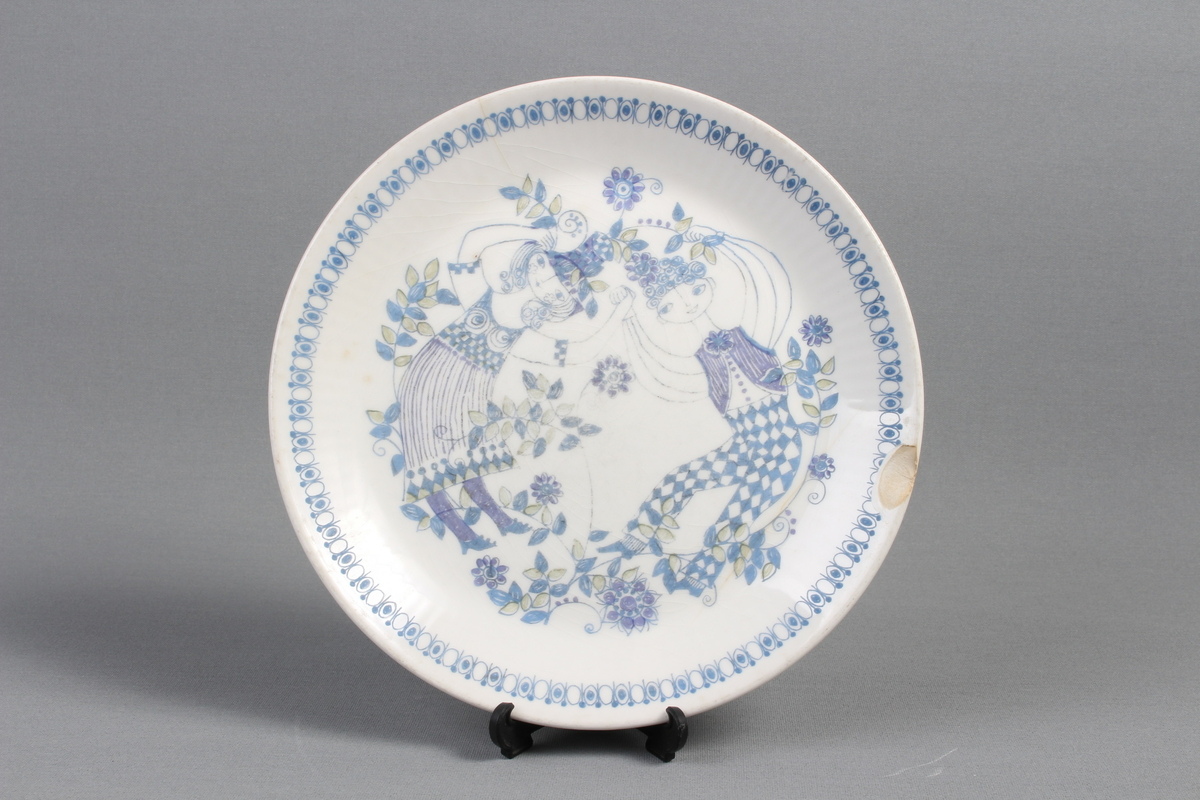 Tallerkene er dekorert med et bilde av en mann  og en kvinne som har blader og blomster på seg og rundt seg. De to menneskene er glade og de danser i blader og blomster.