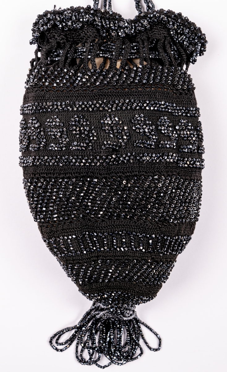 Aftonväska, påsmodell, svart, virkad med inträdda pärlor. Nertill pärlfransar. Väskan bärs i pärlklädda snoddar vilka används för att försluta väskan upptill.
