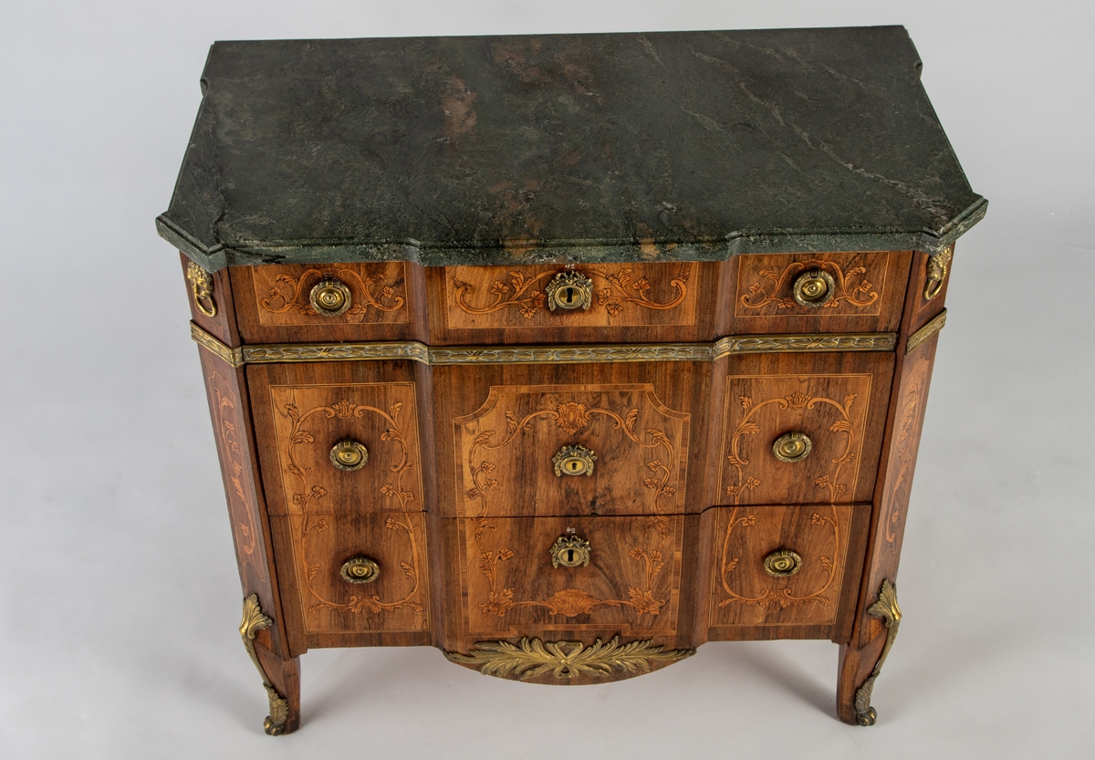 Kommode med knekket front med fremskutt midtparti og avskårne hjørner. Intarsiadekor og bronsebeslag. Profilert marmorplate som følger møbelets form. Forsiden er dekorert med en rekke bronsebeslag: nederst grener med sløyfe, festonger på låsskiltene, rosetter med laurbærkrans på håndtakene, volutter på sidene, palmetter på bena, løvehoder med ring på hjørnene, bronsebånd med laurbærblad under øverste skuff. Intarsiadekor med bladranker.