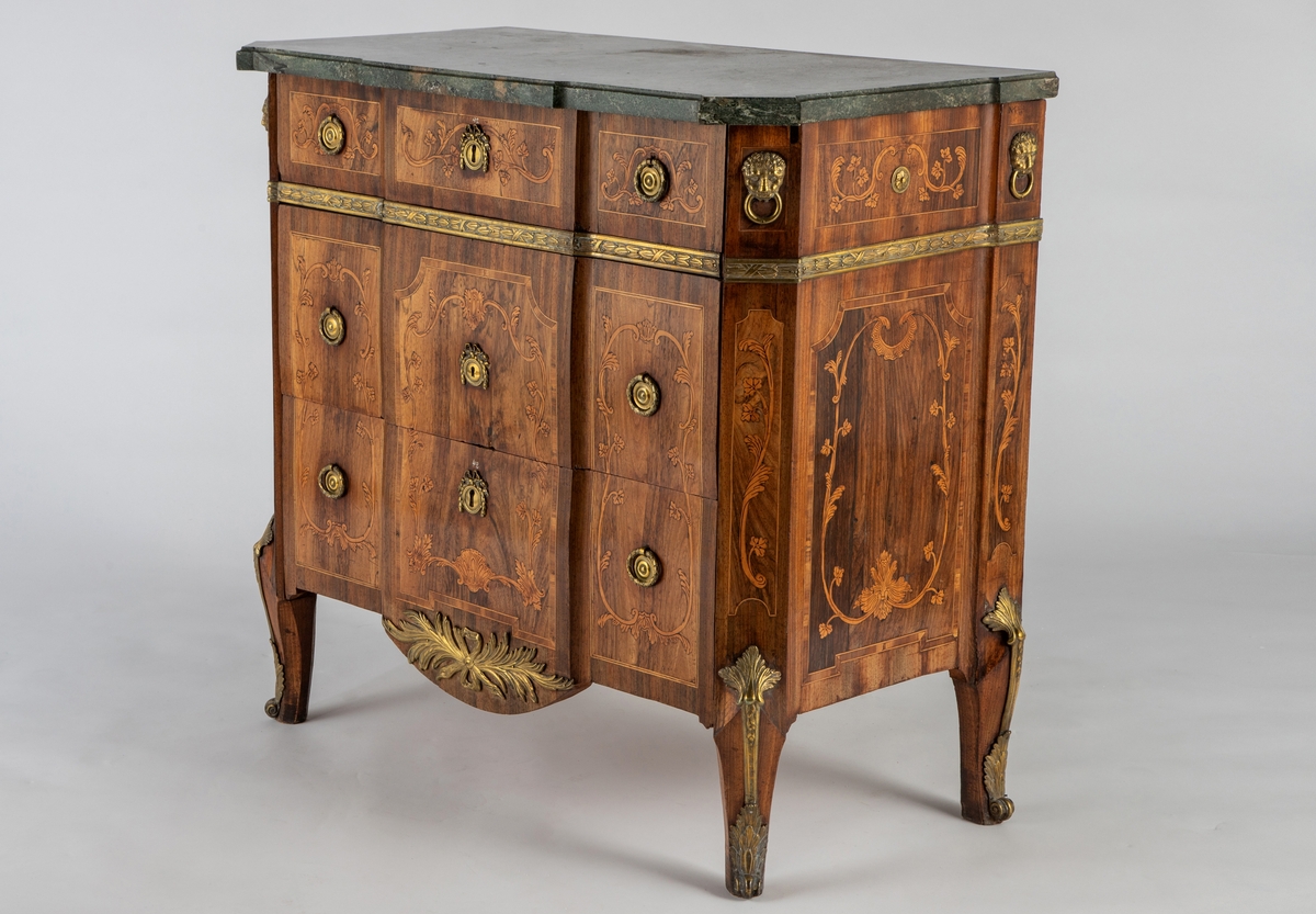Kommode med knekket front med fremskutt midtparti og avskårne hjørner. Intarsiadekor og bronsebeslag. Profilert marmorplate som følger møbelets form. Forsiden er dekorert med en rekke bronsebeslag: nederst grener med sløyfe, festonger på låsskiltene, rosetter med laurbærkrans på håndtakene, volutter på sidene, palmetter på bena, løvehoder med ring på hjørnene, bronsebånd med laurbærblad under øverste skuff. Intarsiadekor med bladranker.