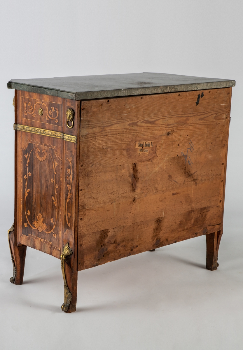 Kommode med knekket front med fremskutt midtparti og avskårne hjørner. Intarsiadekor og bronsebeslag. Profilert marmorplate som følger møbelets form. Forsiden er dekorert med en rekke bronsebeslag: nederst grener med sløyfe, festonger på låsskiltene, rosetter med laurbærkrans på håndtakene, volutter på sidene, palmetter på bena, løvehoder med ring på hjørnene, bronsebånd med laurbærblad under øverste skuff. Intarsiadekor med bladranker.