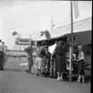 Barn och vuxna vid ett lottstånd i Gränna hamn. Lottståndet har en skylt för Lions. I bakgrunden ligger en färja vid kaj. Sannolikt marknad i Gränna Lions regi. Det är sommar och varmt.