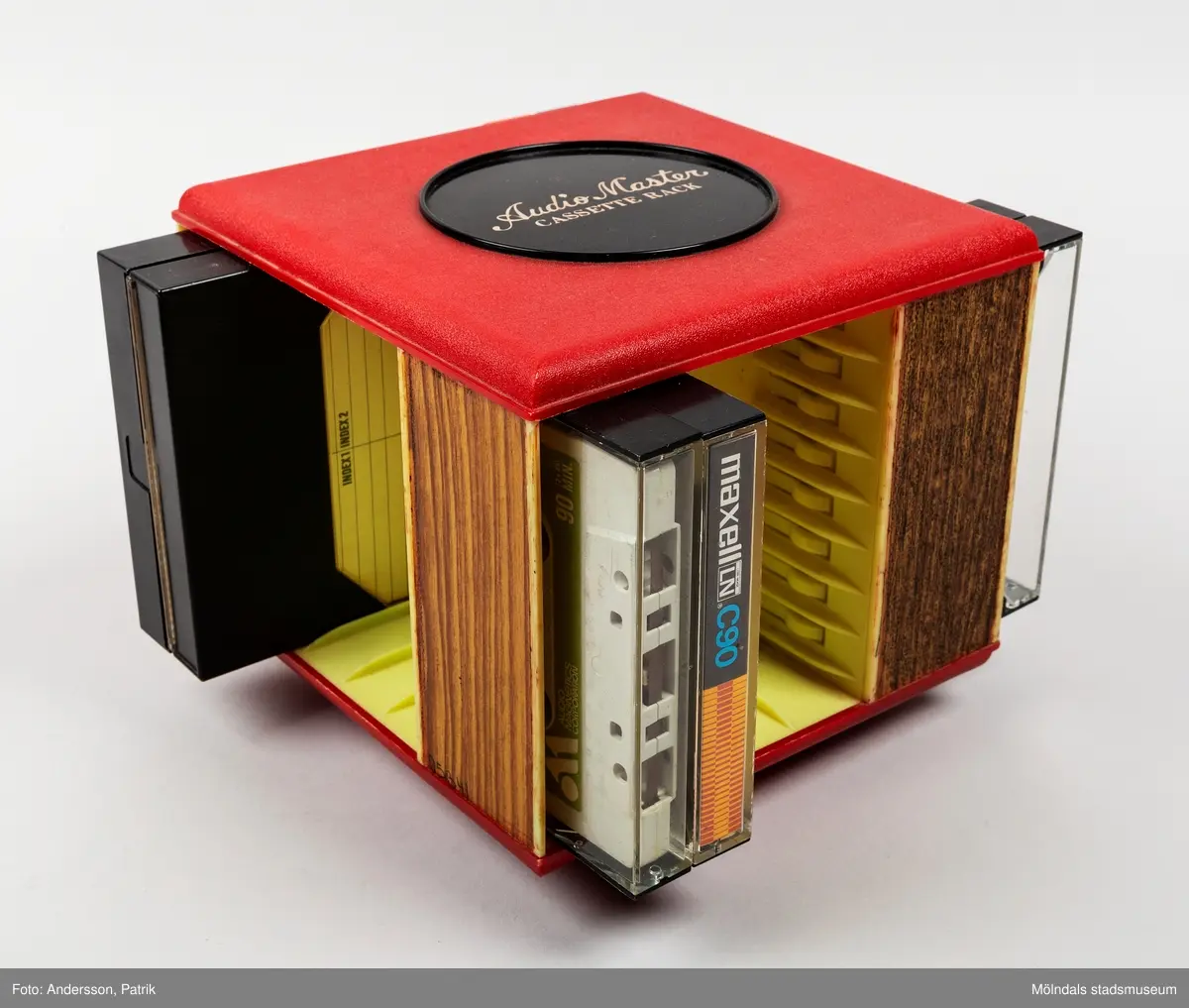 Ett kassettställ av hårdplast för förvaring av kassettband. Stället har en rund fot med manuell snurrfunktion. Kasettställets form är kvadratisk och färgen är röd. Ställets invändiga sidor är ljusgula. Hörnen har en vertikal sida med plast i träimitation. Kassettstället innehåller plats för 20 kassetter. I detta ställ finns 8 kassetter. På ställets ovansida finns en något upphöjd svart cirkel. Inuti cirkelns mitt finns en guldfärgad text med namnet Audio master.
Kassettband introducerades på marknaden 1963 av det nederländska teknikföretaget Philips. De användes för att spela in musik och talinspelningar. Kassettbanden var väldigt populära mellan 1975-1995.