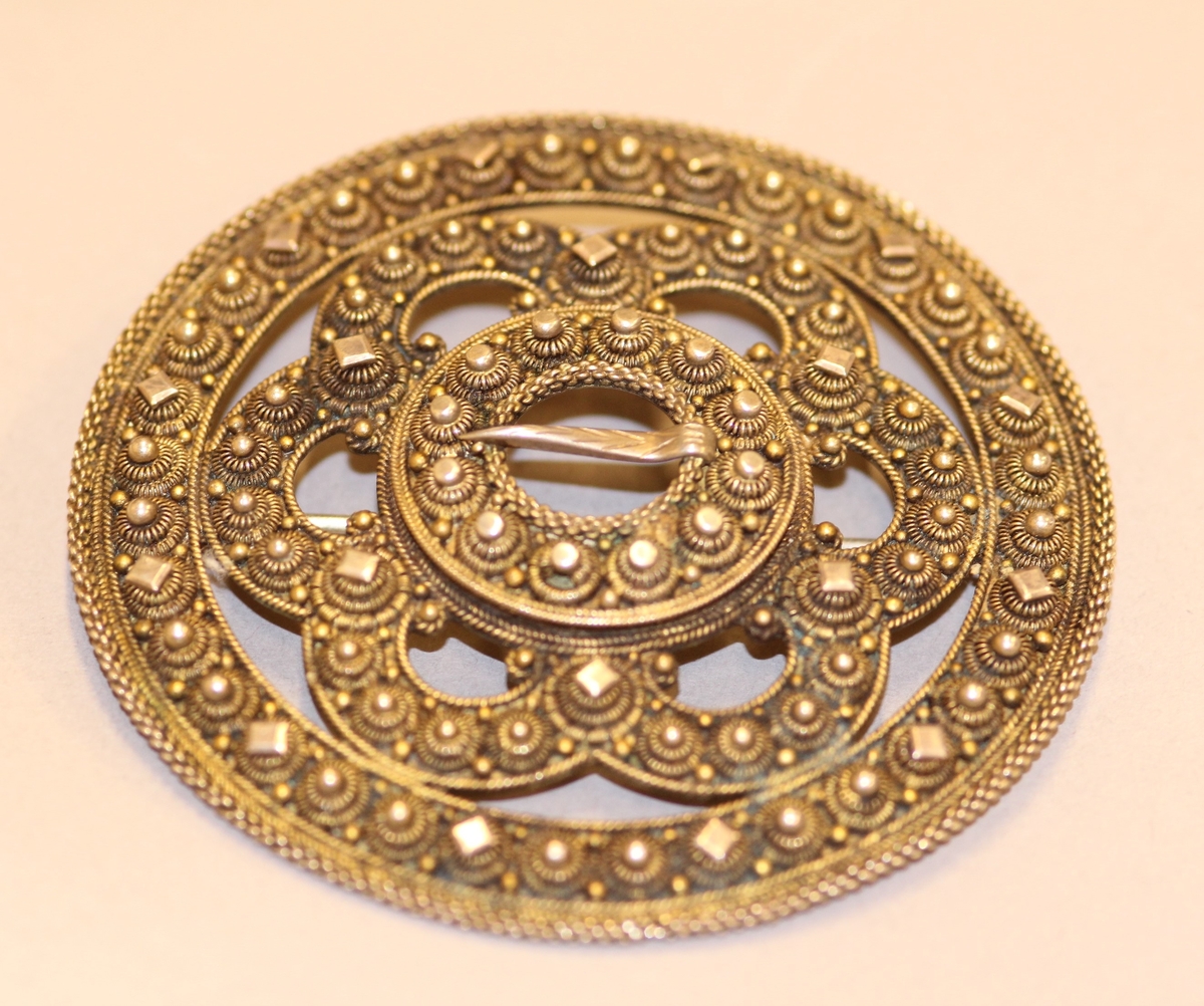 Ein kruse med plate, to krusar med knupp kring ytterste ringen. Inni slangen er det delingskrusar med plater oppå, fire kruser med knupp mellom desse. Rundt spretteholet ei rad med krusar og rund, litt større og flatare knupp. Spretteholet med torn. Sylgja er litt kuva.Nål på baksida.