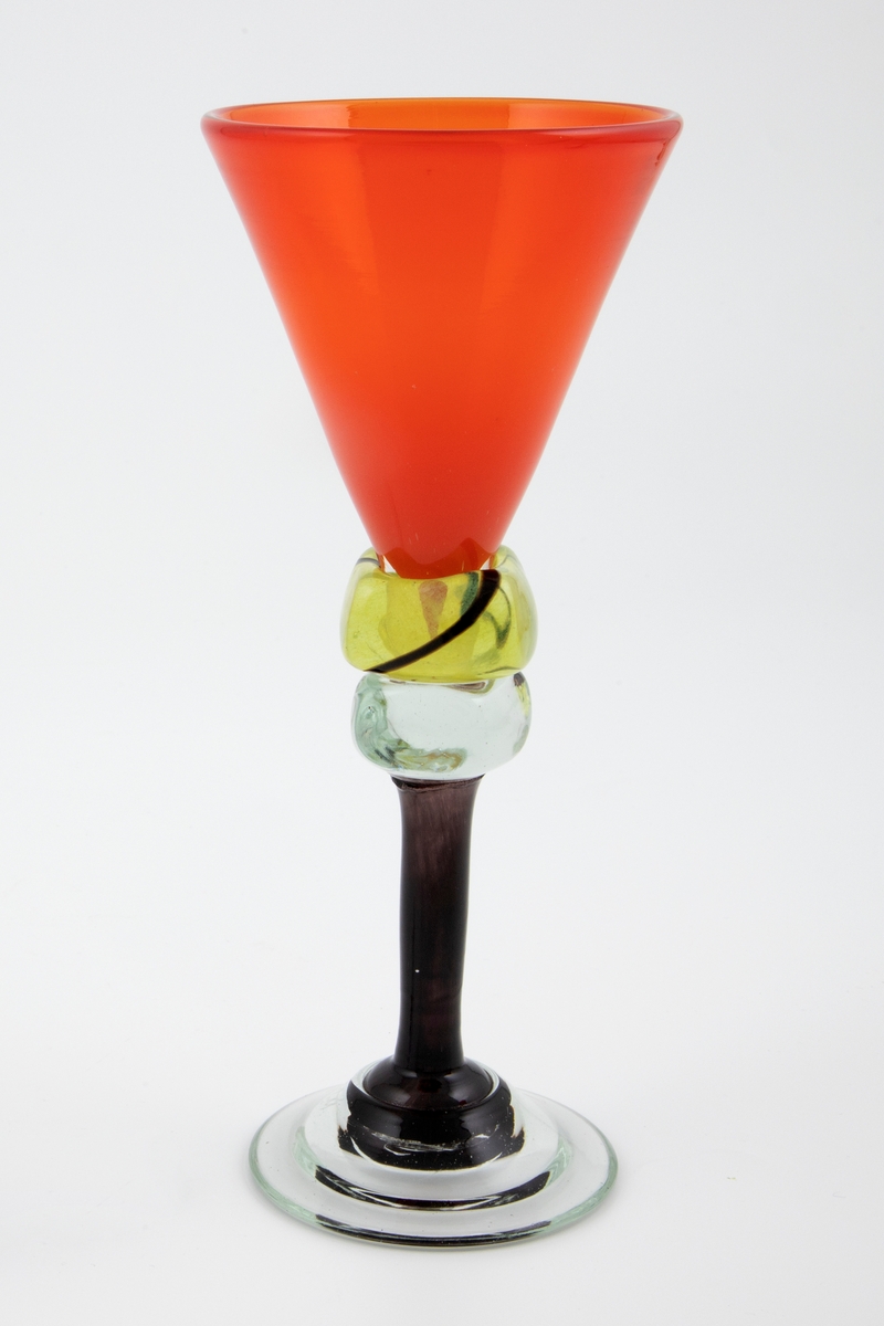 Vinglass i farget glass. Konisk kupa i gjennomskinnelig rødt glass. Sortfarget stett med to terningformede vulster i klart og gult glass, hvorav sistnevnte er dekorert med en bølgende sort linje i spiralform. Sirkulær fot i klart glass, noe hevet, med puntemerke.
