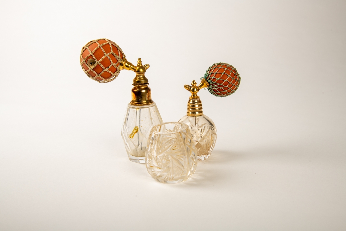 Flakonger i fasettslepet krystall med forgylte topper. Rød gummiballong i grønt trådnett.  Ligger i en såpeeske. Høyde på flakongene: 12 og 10 cm.