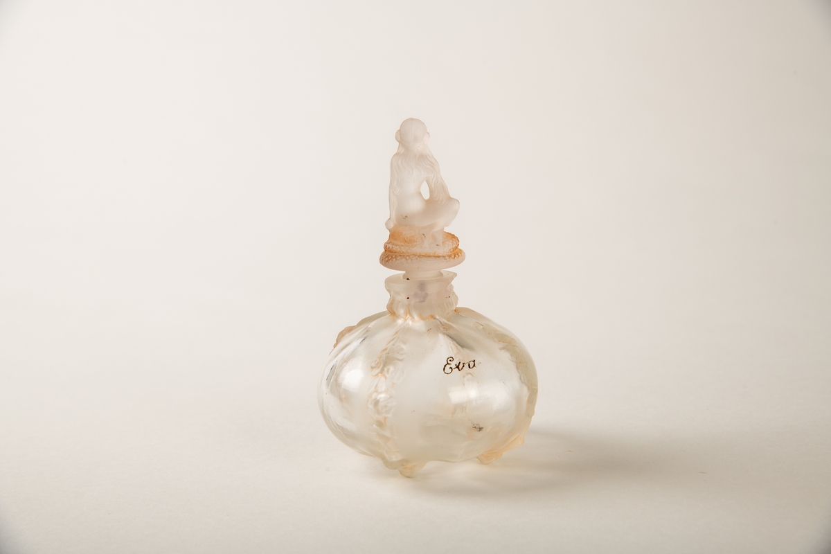 Rund krukke med fire blomsterranker i relieff ned siden. Liten munning med propp, hvorpå en  sittende kvinne på en slange. Inngravert:  Eva.