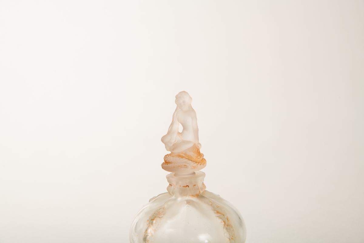 Rund krukke med fire blomsterranker i relieff ned siden. Liten munning med propp, hvorpå en  sittende kvinne på en slange. Inngravert:  Eva.