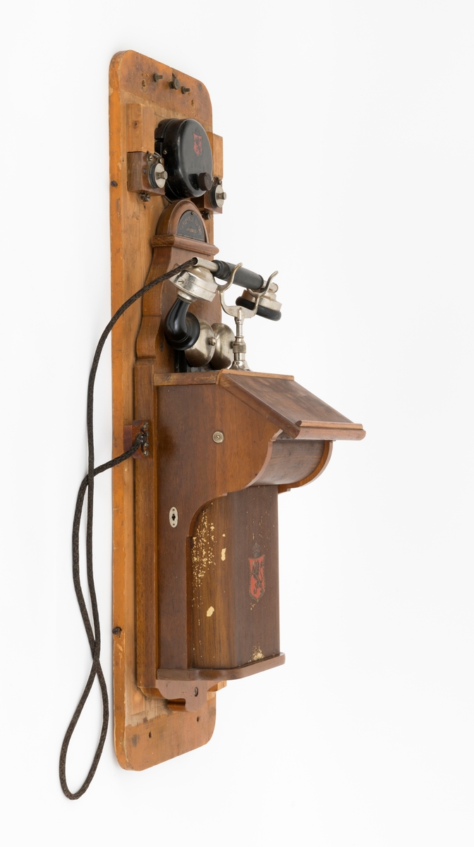 Sveivetelefon, antakelig av type som ble brukt omkring 1. verdenskrig.  Telefonen er montert på ei 1,6 centimeter tjukk treplate.  Apparatet består av ei "kasse" av tre og jernblikk, alt lakkert i edeltreimiterende brunfarge.  I denne delen av apparatet inngår et skrått lokk, en liten skriveklaff, som antakelig skjermer underliggende mekanikk og muligens også batterier.  Ellers har apparatet en forkrommet holder for telefonrøret, som også er forkrommet, men har bakelittkomponenter for håndgrep (midten), øreanlegg og taletrakt.  Apparatet har to omvendt skålformete forkrommete bjeller bak rørholderen, som antakelig gav fra seg klemt når det kom henvendelser per telefon.  Øverst på apparatet er det et slags halvsirkelformet "hode" med informasjon om produsenten.  Her er det også et rødt skjold med en løvefigur med krone over.  Dene logoen finnes også på en svartlakkert, sylindrisk boks på apparatet.  Fra denne boksen er det ledningsforbindelse til stålskiver, som er fastskrudd på begge sider av apparatet. Plata telefonen er montert på har blyantpåskriften «Stor-En-Dammen». Dette antas å være en god indikasjon på at dette apparatet har vært montert i koia ved den nevnte dammen i et av Osensjøens sidevassdrag. Her ble telefonen brukt når fløterne i nedenforliggende deler av vassdraget ønsket å gjøre avtaler som regulerte vanntilførselen i elveløpet på en måte som var optimal ned henblikk på å få tømmeret til å flyte mot Osensjøen.