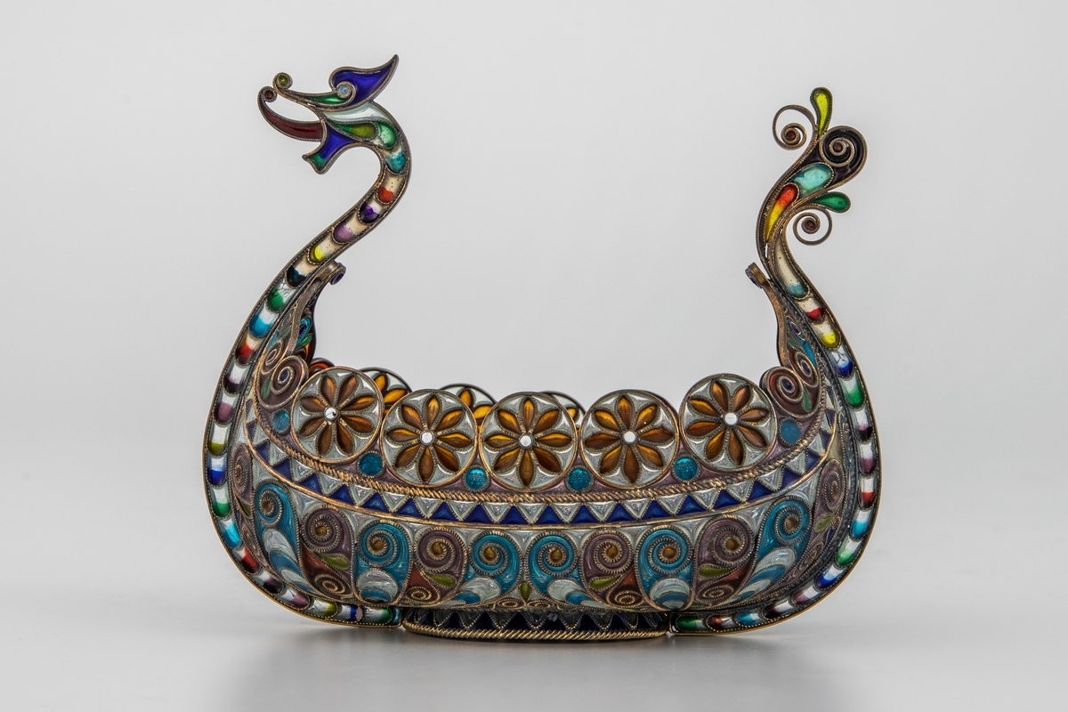 Pyntegjenstand, utformet som et vikingskip, i sølvfiligran og vindusemalje. Oval formet "kjøl" i sølv. Resten av båten er bygget opp av et intrikat filigransarbeid, hvor mellomrommene er fylt med transparent vindusemalje i ulike farger. Filigranstrådene danner rosettformede skjold langs skipets relling, samt voluttornmaenter og s-former på korpus. Forstevnen er utformet som et gapende dragehode, mens akterstaven har avsluttes med blomsterornamenter.