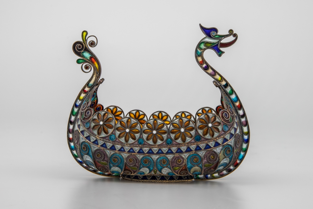 Pyntegjenstand, utformet som et vikingskip, i sølvfiligran og vindusemalje. Oval formet "kjøl" i sølv. Resten av båten er bygget opp av et intrikat filigransarbeid, hvor mellomrommene er fylt med transparent vindusemalje i ulike farger. Filigranstrådene danner rosettformede skjold langs skipets relling, samt voluttornmaenter og s-former på korpus. Forstevnen er utformet som et gapende dragehode, mens akterstaven har avsluttes med blomsterornamenter.