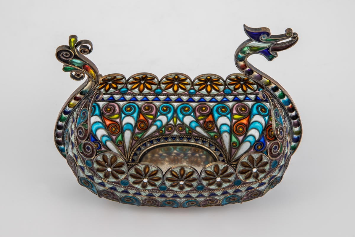 Pyntegjenstand, utformet som et vikingskip, i sølvfiligran og vindusemalje. Oval formet "kjøl" i sølv. Resten av båten er bygget opp av et intrikat filigransarbeid, hvor mellomrommene er fylt med transparent vindusemalje i ulike farger. Filigranstrådene danner rosettformede skjold langs skipets relling, samt voluttornmaenter og s-former på korpus. Forstevnen er utformet som et gapende dragehode, mens akterstaven har avsluttes med blomsterornamenter.