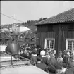 Stor folksamling på Sjögatan nedanför Hallska gården i Gränna. En man med kubb på huvudet sitter i en hästvagn och intervjuas av en man som står nedanför. Mannen som intervjuar är möjligen Herbert Gustafsson. I förgrunden taket av en bil där det sitter en högtalare.