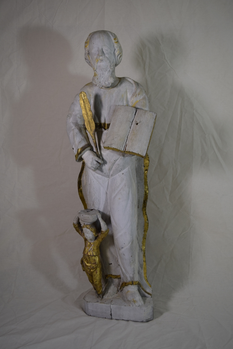 Evangelisten Matteus, stående på høyre ben med engelen ved kneet, oppslått bok på venstre arm. Kappen danner bakgrunn for figuren.