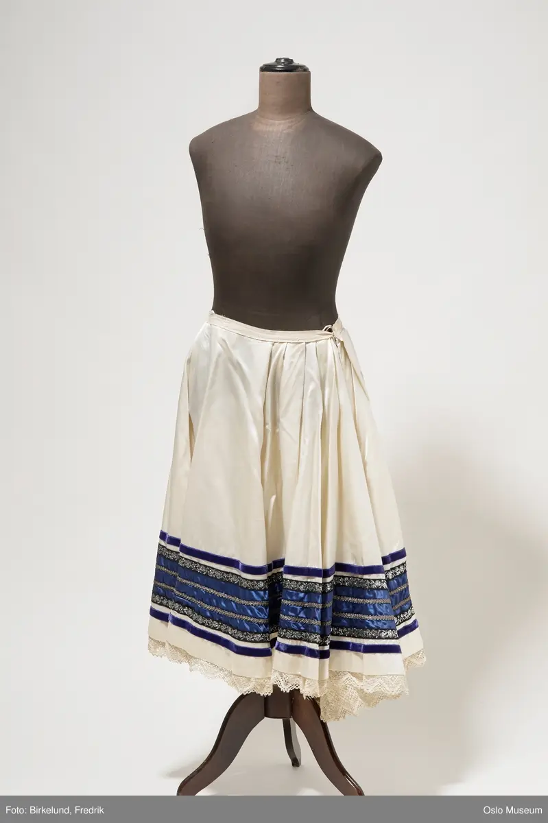 Skjørt i hvit silke. Mot nedre kant av skjørtet er det påsydde bånd i blått, lilla og sølv. Blondekant stikker ut fra under skjørtet. Lukkes med hekte i ryggen.