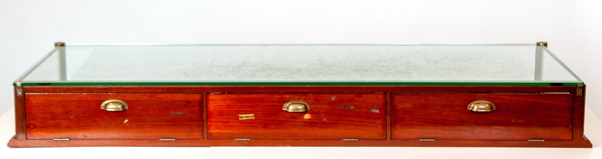 Glaslåda till butiksdisk, JM.56637:1, med rektangulär form och låg höjd. Glaslådan har varit placerad ovanpå butiksdisken. Lådan innehåller tre parallellt liggande lådor under glaset.

Glaslådan består av en låg, profilerad träbotten. Lådans ena långsida och två kortsidor är av glas, liksom ovansidan. Den andra långsidan består av tre nedfällbara träluckor med mässingshandtag och spegelglas på insidan. Glashörnen är sammanfogade med mässingsbeslag. Innanför träluckorna finns uttagbara trälådor med låg kant. Bottnarna är klädda med grön sammet. Under de lösa lådorna är bottenskivan klädd med grön filt. På den mittersta träluckan sitter en klisteretikett med tryckt text: "Engströms UR & OPTIK JÖNKÖPING".