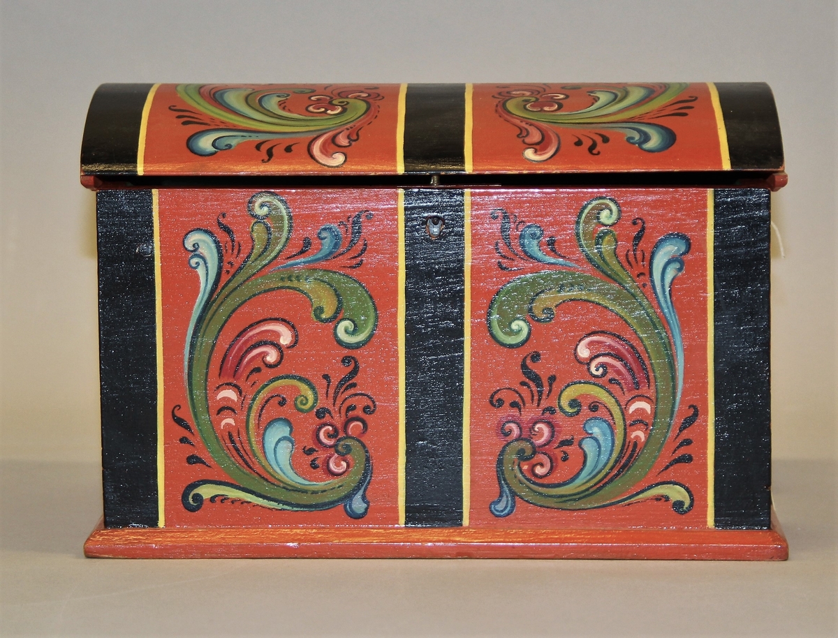 Lita kiste med kuva lok. Rosemåla med tydelege c-krullar. Einsfarga inni, eit lite rom med lok på den eine sida inne i kista.