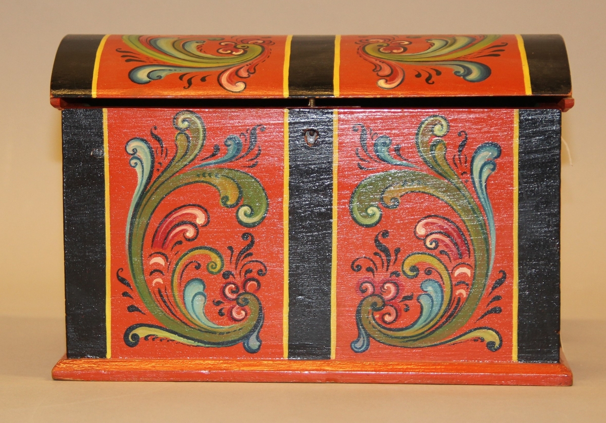 Lita kiste med kuva lok. Rosemåla med tydelege c-krullar. Einsfarga inni, eit lite rom med lok på den eine sida inne i kista.