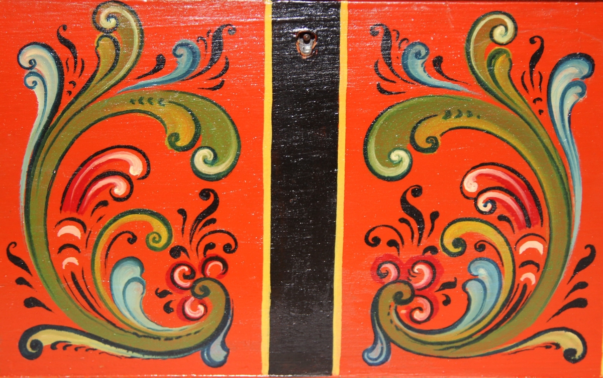 Lita kiste med kuva lok. Rosemåla med tydelege c-krullar. Einsfarga inni, eit lite rom med lok på den eine sida inne i kista.