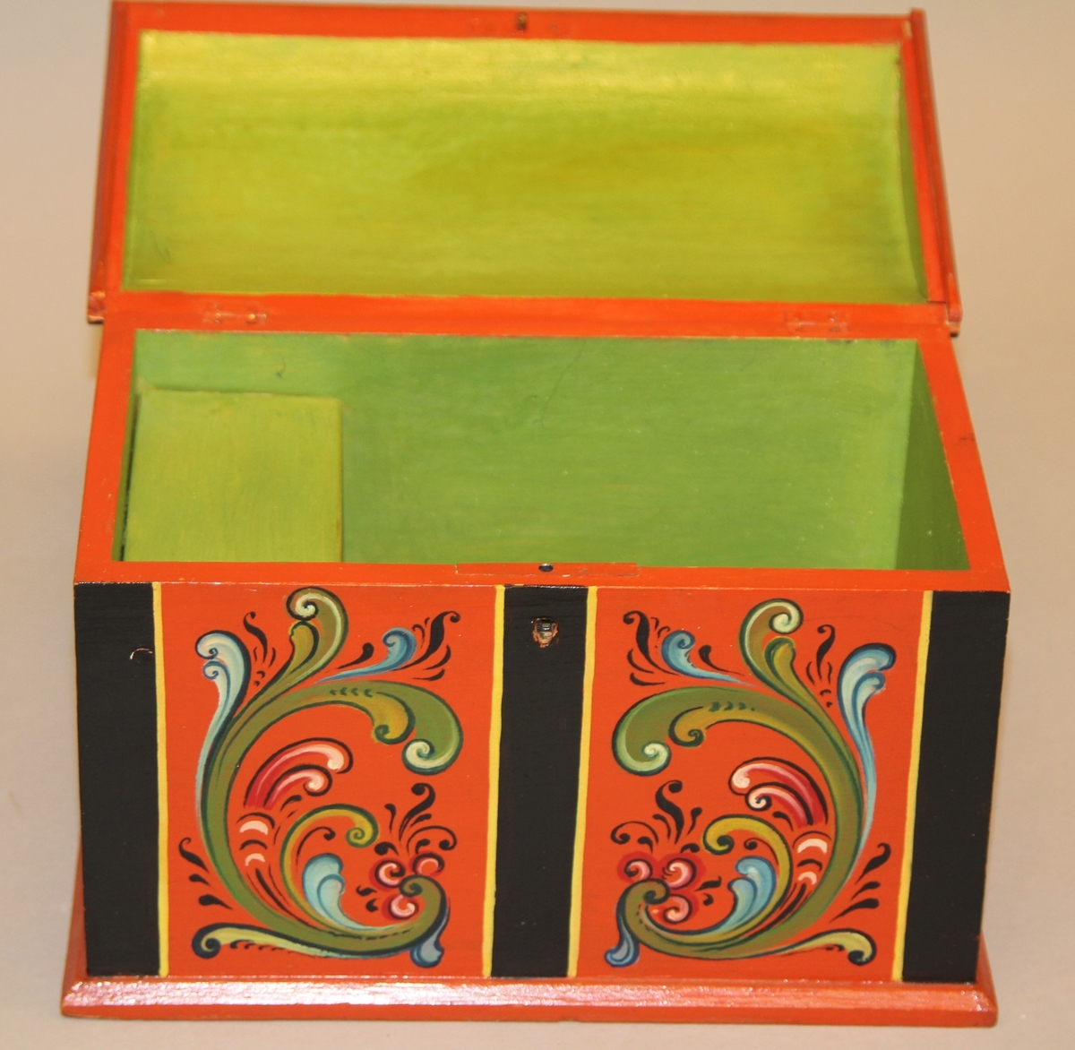 Lita kiste med kuva lok. Rosemåla med tydelege c-krullar. Einsfarga inni, eit lite rom med lok på den eine sida inne i kista.