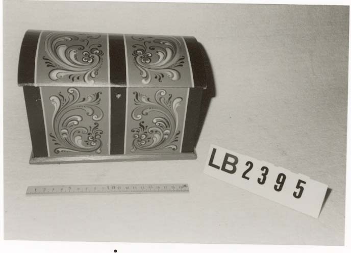 Lita kiste med kuva lok. Rosemåla med tydelege c-krullar. Einsfarga inni, eit lite rom med lok på den eine sida inne i kista.