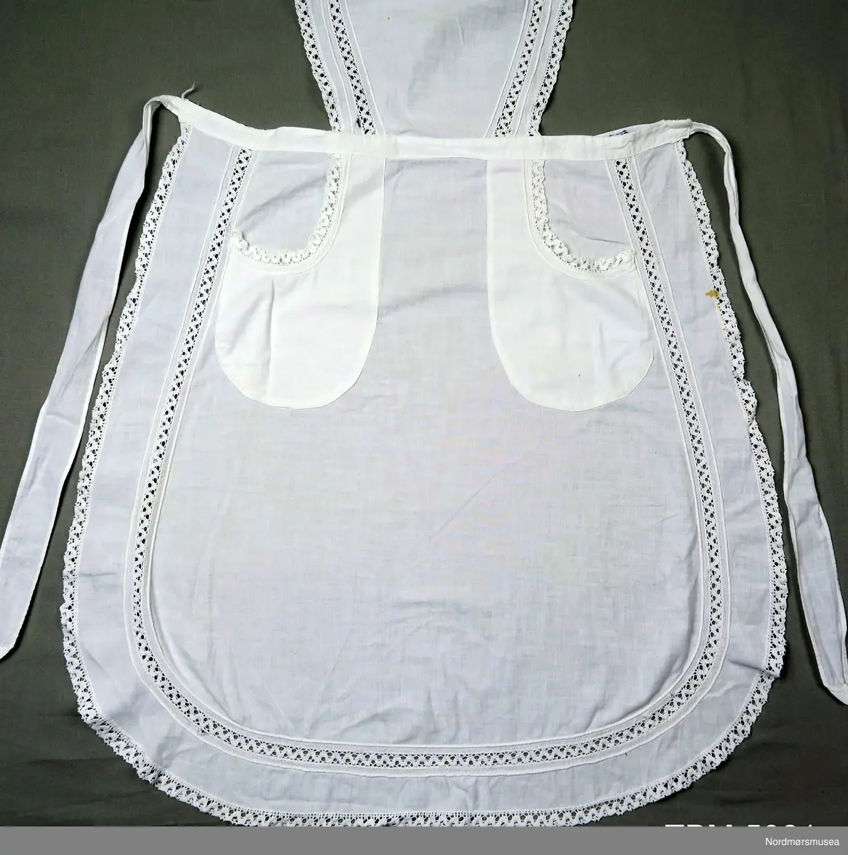 Rundt hele forkleet er det kanter av blonde. Blonder er også innfelt 4 cm inne på smekken og på sjølve forkleet.
To lommmer, også de har blonder. Sjølve forkleet er avrunda nederst.
