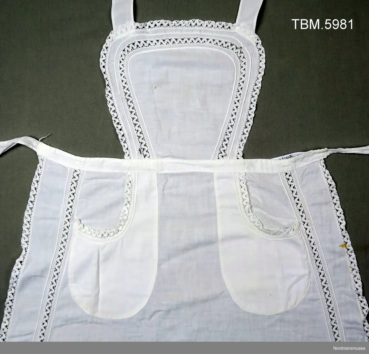 Rundt hele forkleet er det kanter av blonde. Blonder er også innfelt 4 cm inne på smekken og på sjølve forkleet.
To lommmer, også de har blonder. Sjølve forkleet er avrunda nederst.