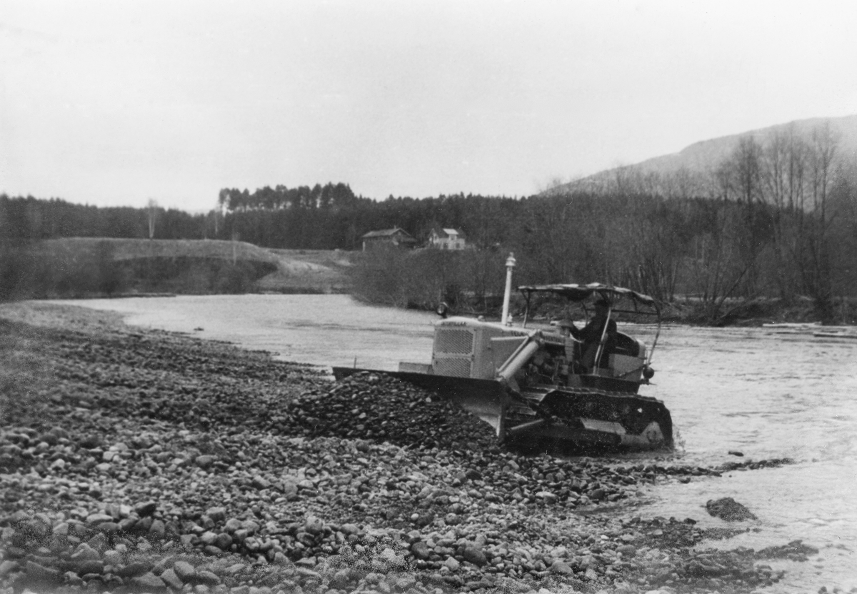 Bulldoser, fotografert i samband med "elverenskingsarbeid" i Skiensvassdraget, antakelig i 1950-åra.  Skiensvassdragets Fellesfløtningsforening leide bulldoser med sikte på fjerning av stein- og grusører i elveløpet i Bøelva i 1953.  På denne måten håpte organisasjonen å kunne redusere problemet med at det la seg hauger eller vaser på slike grunner.  Elverenskingsoppdraget ble lagt ut på anbud, og det var Tele Maskinsentral ved Halvor K. Skjøllaug som fikk oppdraget.  Fotografiet viser beltekjøretøyet som skyver stein- og grusmasser fra elveløpet mot den ene elvebredden.  I det nedenforleggende elveløpet, hvor maskinen har gått, framstår elveløpet som forholdsvis rett og stilleflytende. Samme sesong som den avbildete bulldoseren arbeidet i Bøelva ble det også gjort liknende elverenskingsarbeid i Bygdaråi i Seljord (i nærheten av Lislandbrua), i Flatdal (antakelig Åmotsdalselva ovenfor Flatdalsvatnet) og i Hjartdøla i Hjartdal.  I slutten av 1950-åra påpekte Bø og Nes jakt og fiskelag at Fellesfløtningsforeningen ved denne formen for elverensk kjørte fiskeyngel på land og ødela gyteplassene.  Sportsfiskernes interesseorganisasjon krevde at den ødelagte yngelen ble ersttattet av settefisk, noe Skiensvassdragets Fellesfløtningsforening etter hvert aksepterte. 