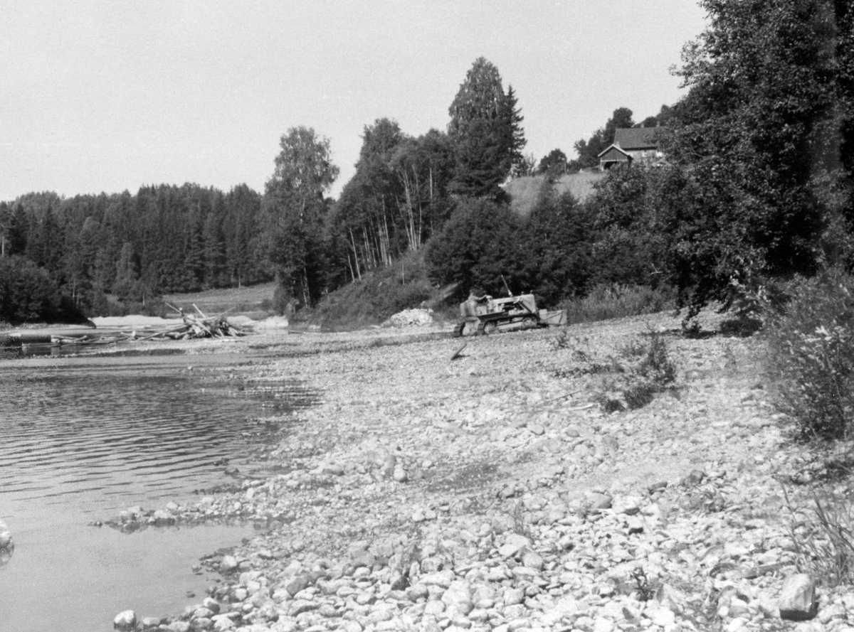 Bulldoser, fotografert i samband med "elverenskingsarbeid" i Skiensvassdraget, antakelig i 1950-åra.  Skiensvassdragets Fellesfløtningsforening leide bulldoser med sikte på fjerning av stein- og grusører i elveløpet i Bøelva i 1953.  På denne måten håpte organisasjonen å kunne redusere problemet med at det la seg hauger eller vaser på slike grunner.  Elverenskingsoppdraget ble lagt ut på anbud, og det var Tele Maskinsentral ved Halvor K. Skjøllaug som fikk oppdraget.  Arbeidet ble videreført de påfølgende sesongene.  Fotografiet viser beltekjøretøyet på ei strand hvor det er skjøvet stein- og grusmasser fra elveløpet mot den ene elvebredden.  Elveløpet hvor maskinen har gått (til venstreb i bildet), framstår som forholdsvis rett og stilleflytende.  Fotografiet skal være tatt i nærheten av et sted i Bø som heter Leikvang.  Samme sesong som bulldoserarbeidet i Bøelva startet ble det også gjort liknende elverenskingstiltak i Bygdaråi i Seljord (i nærheten av Lislandbrua), i Flatdal (antakelig Åmotsdalselva ovenfor Flatdalsvatnet) og i Hjartdøla i Hjartdal.  I slutten av 1950-åra påpekte Bø og Nes jakt og fiskelag at Fellesfløtningsforeningen ved denne formen for elverensk kjørte fiskeyngel på land og ødela gyteplassene.  Sportsfiskernes interesseorganisasjon krevde at den ødelagte yngelen ble ersttattet av settefisk, noe Skiensvassdragets Fellesfløtningsforening etter hvert aksepterte. 