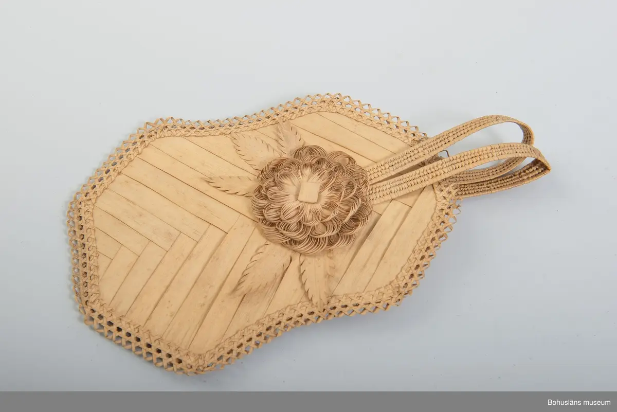 Ur handskrivna katalogen 1957-1958:
Solfjäder av trä, Amerika
Mått: 30 x 18 cm; av bast och tunna träfibrer; en mångsid. platta m. runda hörn, jämte handt. Hel.

Ur handskriven katalog över Uddevalla - Musei Historiska - samlingar: Utländska föremål = De etnografiska föremålen upprättad  år 1916 av Knut Adrian Andersson, Intendent från 1 Oktober 1915:
Solfjäder (Fläkt) av trä (     ), gjord av infödingar å St Barthélèmy 1840talet. Skänkt av fröken Anna Craelius i Uddevalla.

Troligen gjort av strå av någon sorts palmblad.