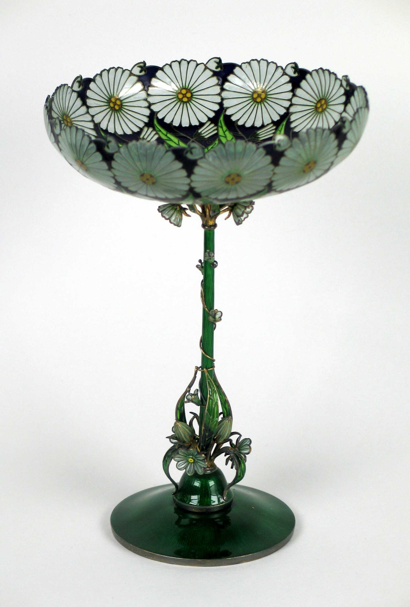 Skålformet vase med lang, tynn stett og opphøyet fotstykke. Foten er laget i sølv og grønn emalje. Fra denne slynger det seg blomster, knopper og bladverk oppover stetten. Oppunder den flate glasskålen er det tre blomster, knopper og blad. I selve skålen er det innerst en rosett med gullkule i midten. Videre er det samme mønsteret gjentatt på mørk blå bunn; to grønne blad, en knopp og øverst en utsprungen margeritt.