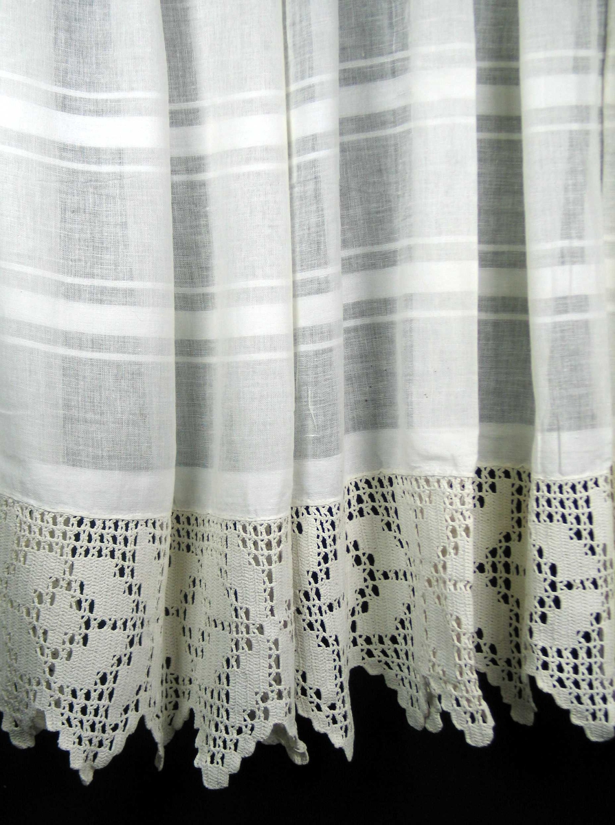 Hvit gardin med striper og heklet blonde sydd på for hånd. Løpegang og ellers faldet med maskin.