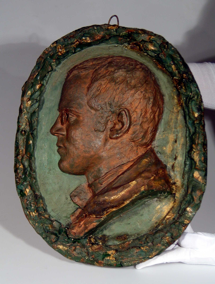 Ovalt gipsrelieff av en mann i profil. Krans rundt.