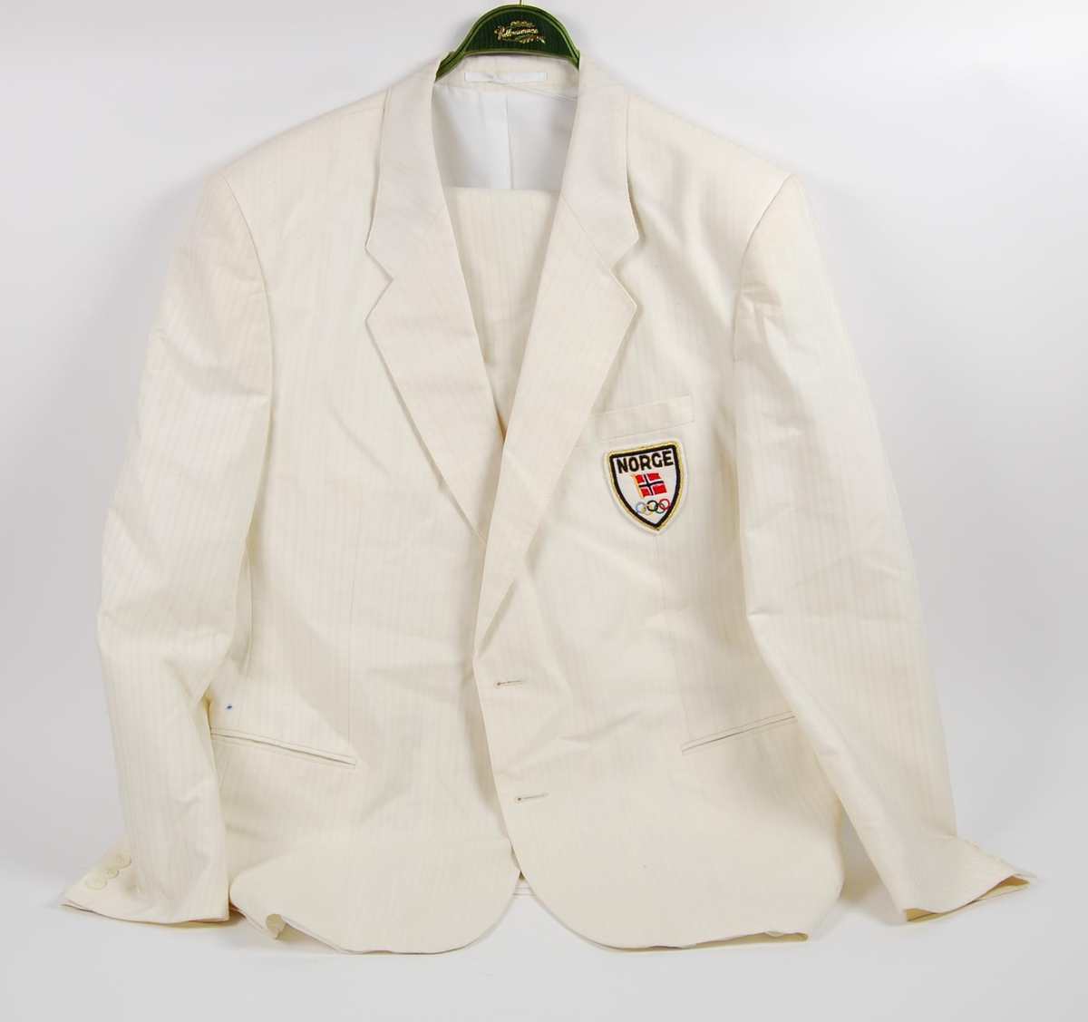 Hvit dress bestående av jakke og bukse. På jakka er det påsydd et merke med de olympiske ringene, norske flagget og innskriften: NORGE. 