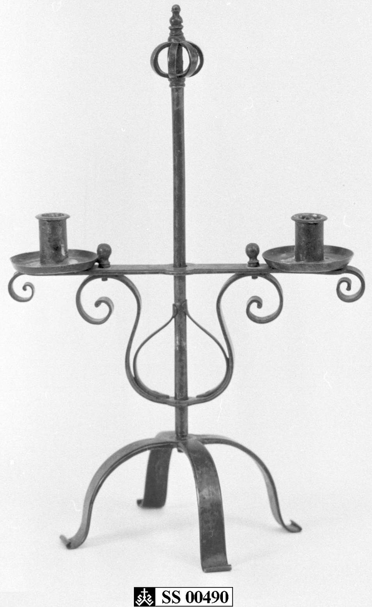 Staken er sammensatt av smidde deler, og består av en firbeint fot med en stang, toppet av en krone. På stangen er en lysarm med to lysholdere. Lysarmen er justerbar i høyden.