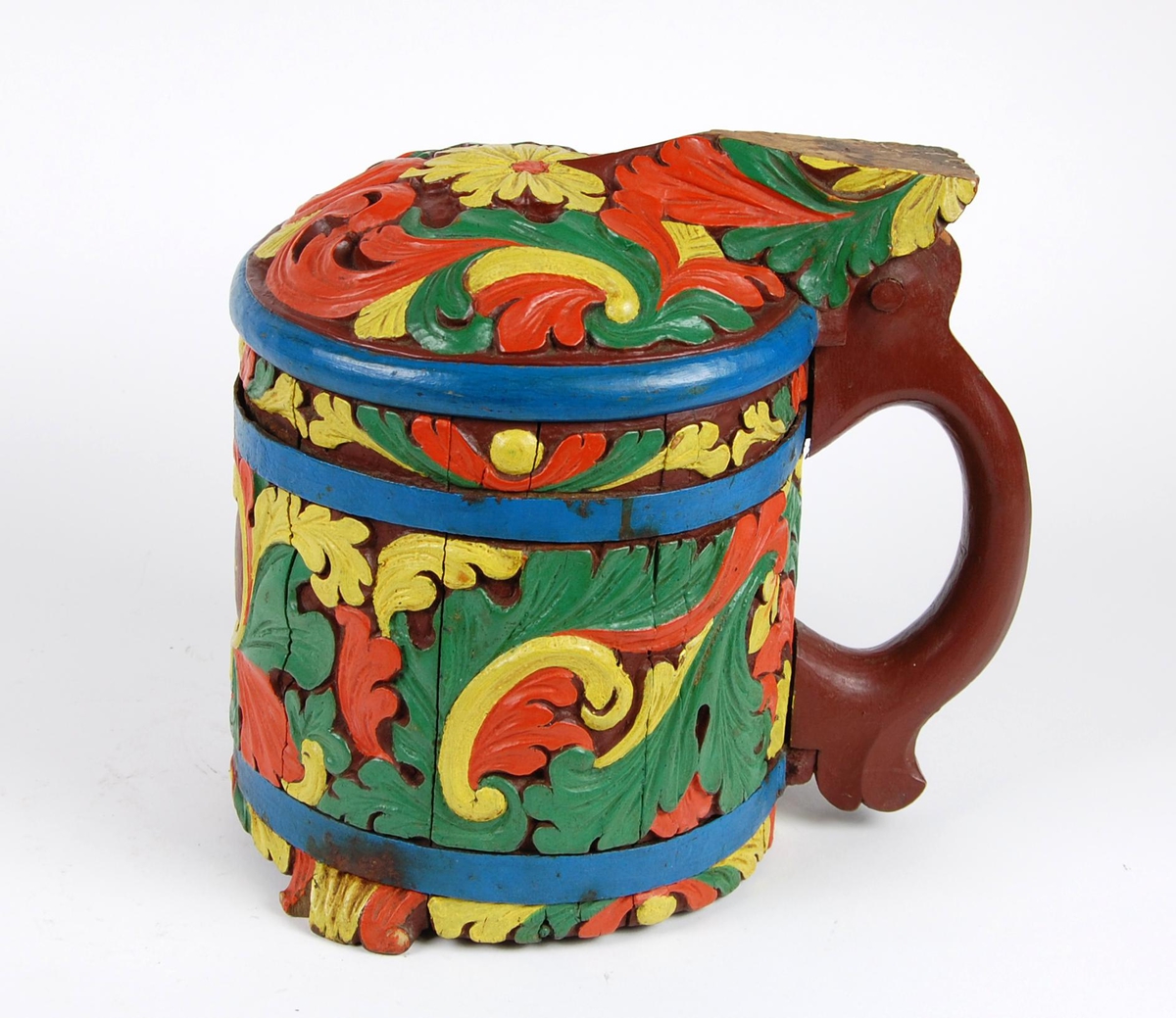 Kanne av staver. Lokk med gule, grønne og røde blader, blåmalt kant. Korpus med en blåmalt metallgjord oppe og nede, gule, grønne og røde akantusblader, brunmalt hjerteformet felt med innskriften 1865. Brunmalt hank. Tre røde og gule føtter.