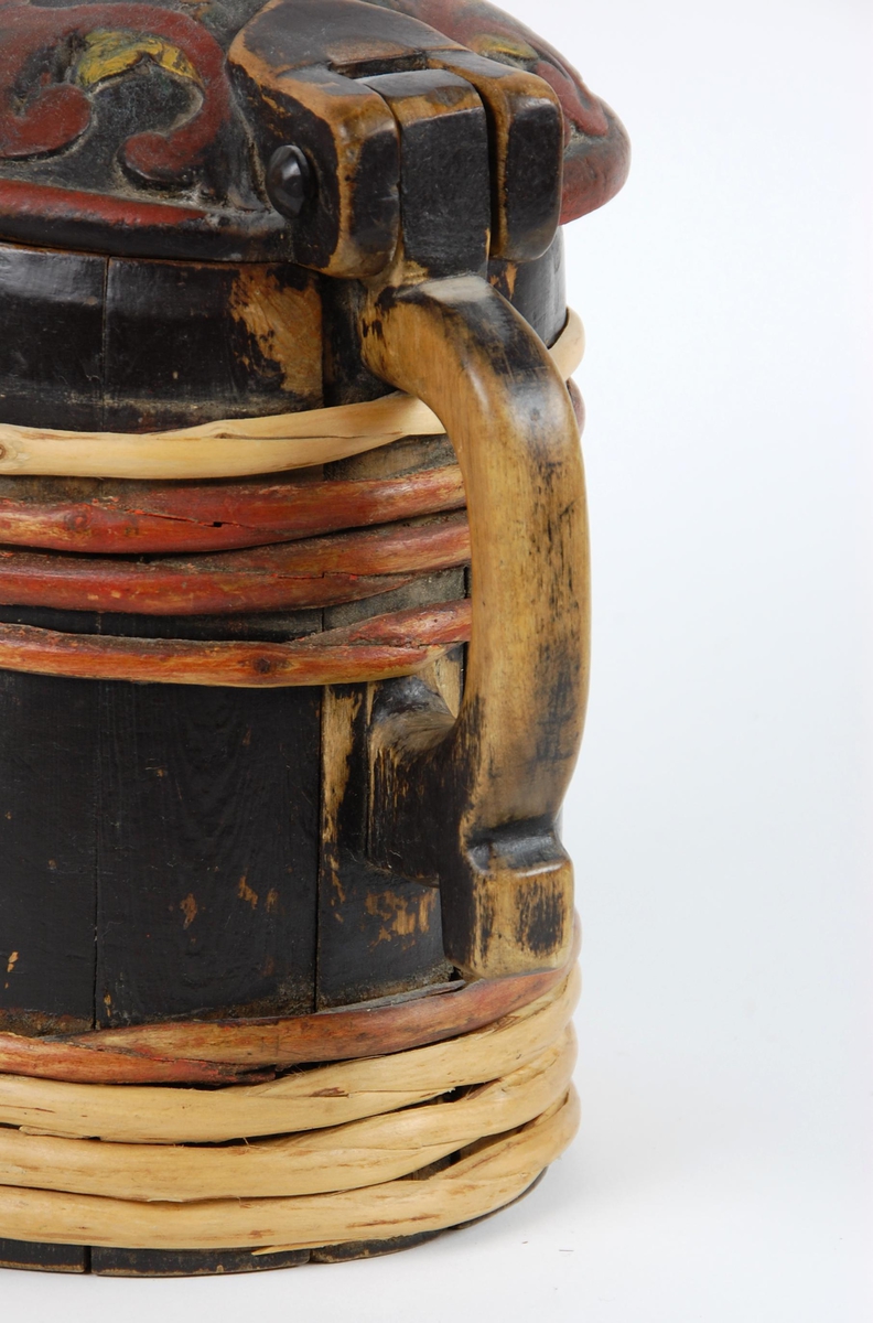 Kanne av trestaver. Lokk med rødt, gult og grønt akantusskurd. Korpus med fire gjorder oppe og nede, fire av gjordene er av nyere dato, sotmalet. hanken er skåret ut av en stav.