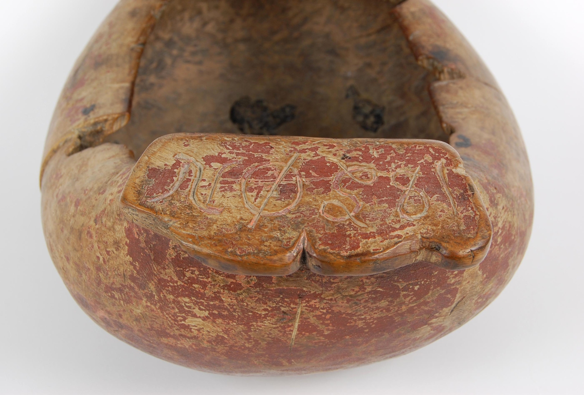 Trøys av bjørk. Oval form. Utskåret mannshode med munnåpning. Håndtak med hakkformete utskjæringer og innskrift. Svarte bekklumper i bunnen.